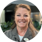 A woman with long hair is smiling at the camera, wearing a gray shirt and a badge. The background features indoor plants and blurred furnishings.