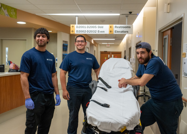 Three men in navy blue shirts stand with a stretcher in a hospital hallway. They are smiling and wearing blue gloves. A directional sign above points to restrooms and elevators. The setting is well-lit with a nurse's station visible in the background.