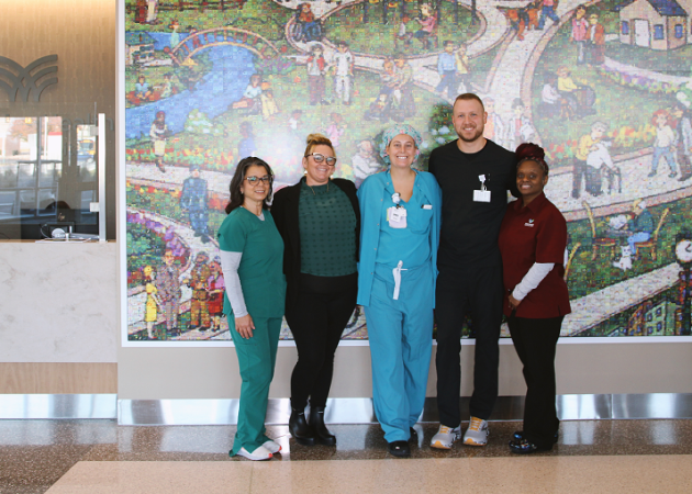 Five healthcare workers stand together smiling in front of a vibrant mural depicting a park scene. Behind them is a hospital reception desk. They wear a mix of scrubs and casual attire, showcasing a diverse team in a healthcare setting.