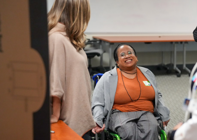 A person in a wheelchair, wearing glasses and an orange top, smiles while talking to another person with long hair in a beige sweater. They are indoors, in a room with tables and a whiteboard.