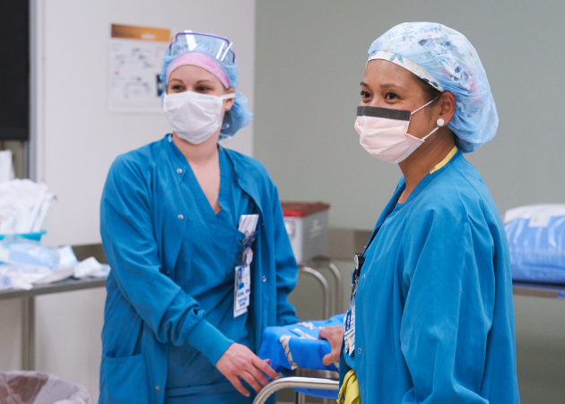 Two medical professionals in scrubs and masks are in a hospital setting. One is holding a package by a metal trolley. Both are wearing hair covers and looking in the same direction, appearing focused and attentive.