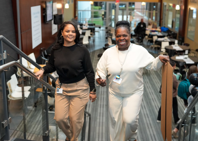 Two women smiling and walking up a staircase indoors. The woman on the left wears a black top and beige pants, while the woman on the right wears an all-white outfit and holds a folded piece of cardboard. Behind them are tables and people sitting.
