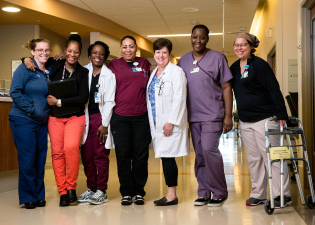 A group of seven healthcare professionals of diverse gender and race stand together in a hospital corridor, smiling. They are wearing various medical uniforms, including scrubs and a lab coat. One person is using a walker.