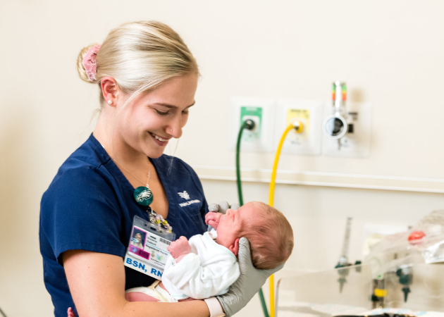 A nurse in blue scrubs is smiling while holding a newborn baby wrapped in a white blanket. They are in a hospital room with medical equipment visible in the background.