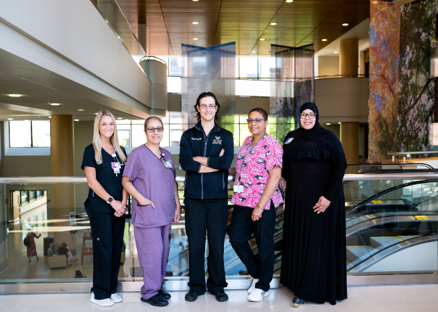 A group of five healthcare professionals in a hospital setting. They are standing in a modern, spacious interior with escalators visible in the background. They are smiling and wearing uniforms in various colors.