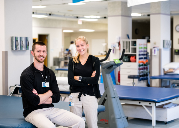 Two smiling healthcare professionals wearing black tops and beige pants stand in a bright rehabilitation facility. One sits on a treatment table, while the other stands with arms crossed. The room contains therapy equipment like a treadmill and weights.