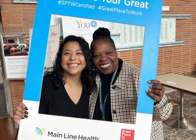 Two smiling women are holding a large blue picture frame with hashtags #GPTWCertified and #GreatPlaceToWork. The frame has the "Main Line Health" and "Great Place To Work" logos at the bottom. They are indoors with a brochure display behind them.