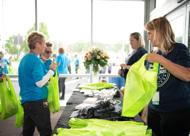 People at an event registration table distribute bright green bags. Participants wear blue event shirts, and there is a bouquet of flowers on the table. The setting is bright, with large windows in the background.
