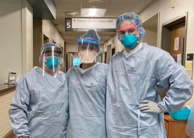 Three healthcare workers in full protective gear, including gowns, masks, face shields, and gloves, stand together in a hospital corridor. A sign indicates "Patient Rooms." The scene reflects a clinical, supportive environment.