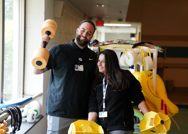 Two people smiling, holding foam dumbbells in a therapy center. One man raises a dumbbell in one hand, while a woman holds another, surrounded by various therapy equipment. They appear cheerful, engaged in a healthcare setting.