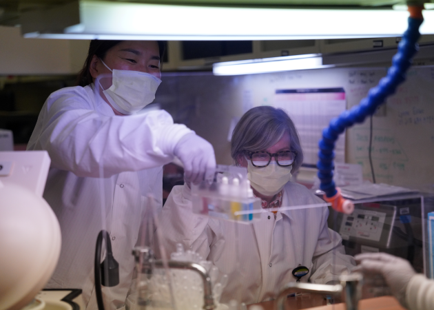 Two scientists in lab coats and masks work in a laboratory. One stands, holding a rack of test tubes, while the other sits, observing. They are surrounded by lab equipment and work under bright overhead lighting.