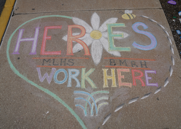 Colorful chalk drawing on concrete reads "Heroes Work Here" in a heart shape. A daisy and bee embellish the text, with "MLHS" and "BMRH" included. Dotted lines and decorative elements enhance the design.