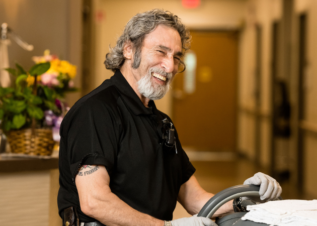 Smiling man with gray hair and beard, wearing a black shirt and gloves, is cleaning with a cart in a hallway. A basket of flowers is visible in the background.