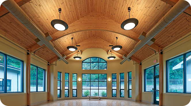 A spacious room with a high, wooden vaulted ceiling and large windows. Round overhead lights are evenly spaced, and the room has light wooden floors. Windows provide a view of greenery outside.