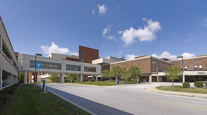Outside view of a hospital building with a sky bridge, multiple wings, and a main entrance. The structure has a mix of brick and light-colored exteriors, surrounded by neatly trimmed lawns and trees. A blue sky with scattered clouds is above.