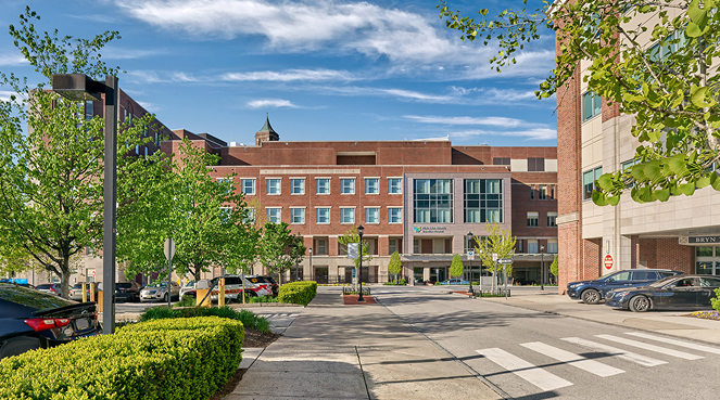 Street view of a hospital entrance with a multistory brick building. Green trees and hedges line the pathway, and parked cars are visible. A crosswalk leads to the entrance under a blue sky with scattered clouds.