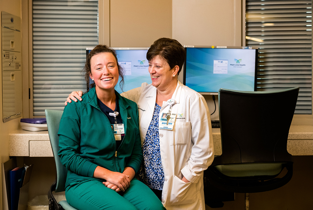 Two healthcare professionals, one sitting and one standing, smiling together in a clinical setting. They wear ID badges and are in front of computer monitors, with medical cabinets and blinds in the background.