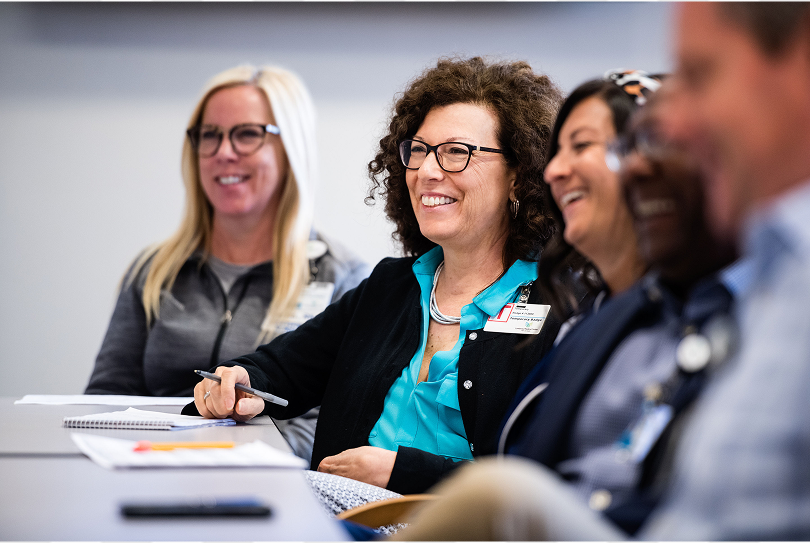 A group of people sitting at a table in a meeting. The focus is on a smiling woman with curly hair, glasses, and a badge. Others are seated beside her, also smiling. Note-taking materials are on the table.