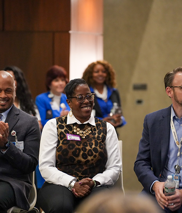 A diverse group of people seated and smiling in a conference setting. The central woman wears a leopard-print vest. Others are in professional attire. They appear to be engaged and attentive, with more people blurred in the background.