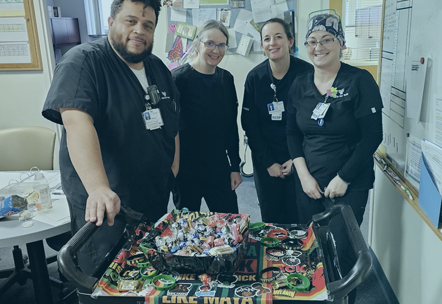 Four healthcare workers in scrubs are smiling and standing behind a cart filled with a variety of candy. The cart is decorated with colorful posters. A bulletin board with papers and notes is visible in the background.