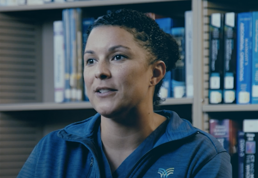 A person with curly hair is speaking, wearing a blue jacket with a logo. They are seated in front of shelves filled with books.