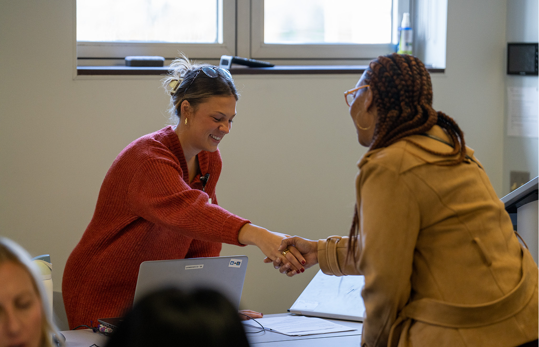 Two women shake hands across a table in a bright room. One is wearing a red sweater and sitting in front of a laptop, while the other is standing in a tan coat. They are smiling and engaged in conversation.