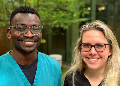 Two people smiling outdoors. The person on the left wears glasses and turquoise scrubs, while the person on the right has long blonde hair, wears glasses and black scrubs. They stand in front of greenery and a building with large windows.
