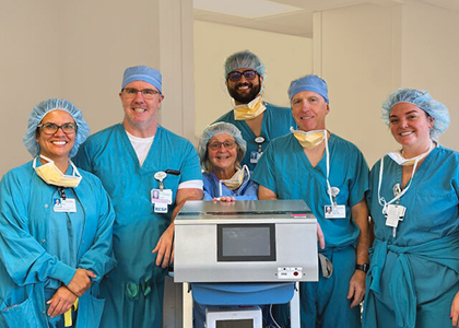 A group of six healthcare professionals wearing blue surgical scrubs and caps stand around a medical device on a cart. They are smiling at the camera in a brightly lit hospital room.