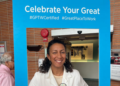 Smiling person with curly hair wearing a white coat stands in a blue frame with text "Celebrate Your Great #GPTWCertified #GreatPlaceToWork." Brick wall and a small exhibition are visible in the background.