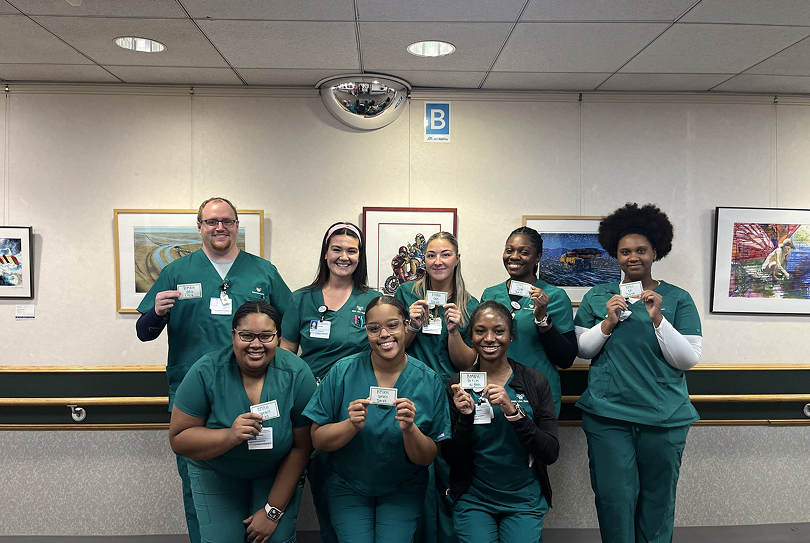 Eight healthcare workers in green scrubs stand indoors, smiling and holding up cards. They pose in front of a wall with framed artwork and a convex security mirror above them.