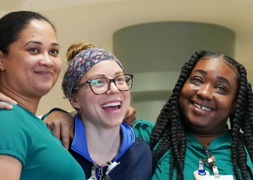 Three women in medical scrubs smile and pose together. One wears glasses and a colorful headband, while the others have their hair down and in braids. They appear happy and friendly in a bright indoor setting.