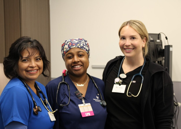 Three healthcare professionals wearing scrubs and stethoscopes smile at the camera in a clinical setting, standing closely together.