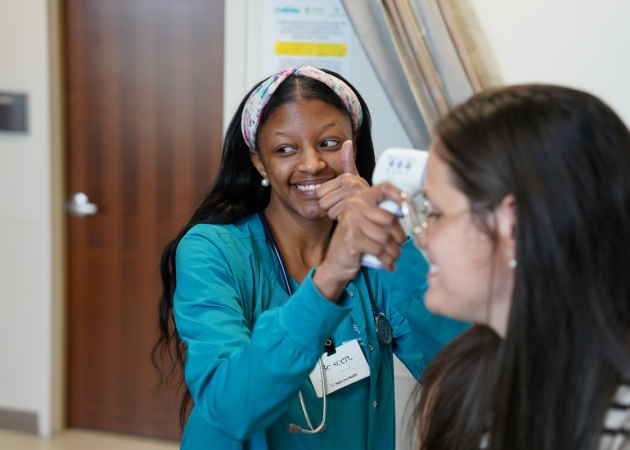 A healthcare worker in teal scrubs smiles while using a medical device to check the forehead of a patient with long brown hair in a clinical setting.