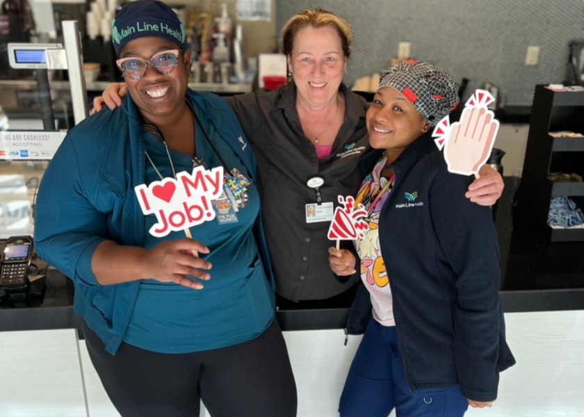 Three smiling women stand together indoors. Two wear medical scrubs, and one wears business attire. They hold cheerful signs, including one that says "I ♥ My Job!", celebrating in a bright, friendly setting.