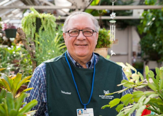 A smiling older man wearing glasses, a plaid shirt, and a green vest with “Volunteer” written on it stands in a greenhouse surrounded by various green plants. He wears an ID badge on a blue lanyard.