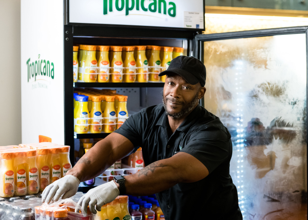 A man in a black shirt, cap, and gloves stocks bottles of Tropicana juice in a refrigerated display, smiling at the camera. Shelves filled with orange juice bottles are visible behind him.