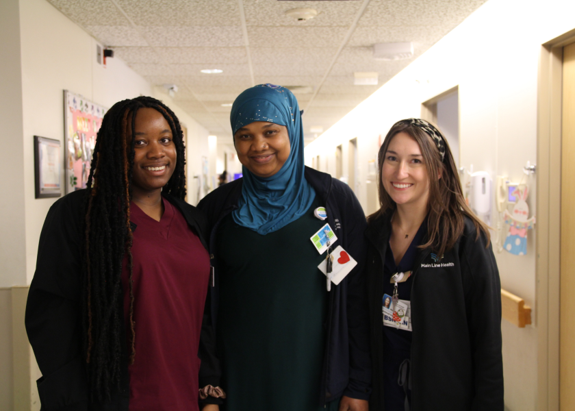 Three healthcare workers stand side by side in a hospital hallway, smiling at the camera. They are wearing scrubs and ID badges; one wears a blue headscarf. The hallway is bright, with doors and decorations visible.