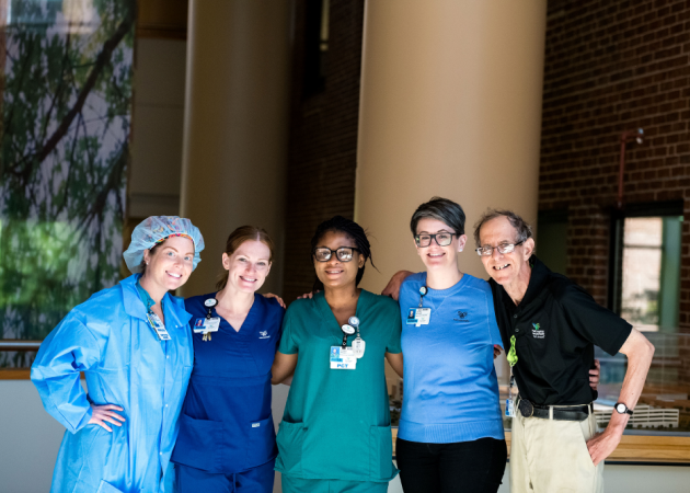 Five healthcare professionals stand together indoors, smiling with their arms around each other, wearing scrubs and uniforms, against a backdrop of large windows and brick walls.