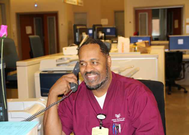 A man in burgundy medical scrubs sits at a desk in a healthcare facility, smiling and talking on the phone. Computer monitors and office supplies are visible in the background.