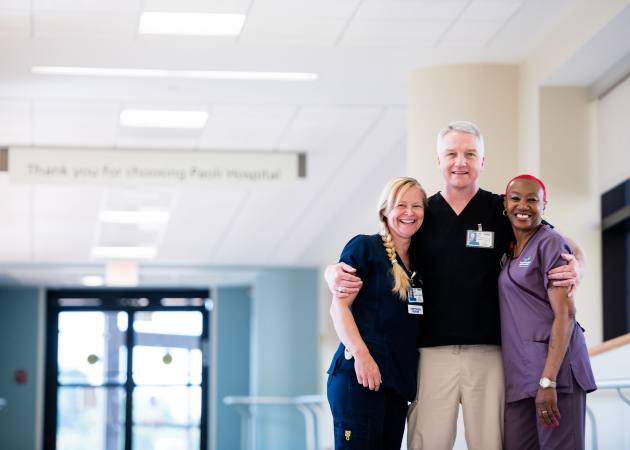 Three smiling healthcare professionals, two women and one man, stand together in a hospital hallway, wearing scrubs and name badges. They have arms around each other, suggesting camaraderie and teamwork.