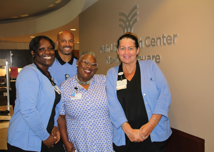 Four smiling people wearing blue and black work attire stand together in front of a wall sign that reads “Main Line Health Center Newtown Square.” They appear to be staff members posing for a group photo.