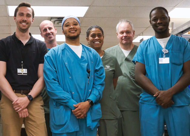 Six healthcare professionals, including men and women, stand together smiling in a hospital setting. Four wear scrubs, while two are in casual work attire. The background shows a ceiling with fluorescent lights.