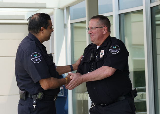 Two police officers in uniform stand outside a building, smiling and shaking hands in a friendly manner. One officer has dark hair, while the other wears glasses. They appear to be having a positive interaction.