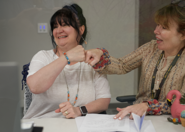 Two women sit at a desk, smiling and giving each other a fist bump. Papers and a pink flamingo figurine are on the table in front of them. One woman wears a white shirt and beaded jewelry; the other wears a patterned blouse.