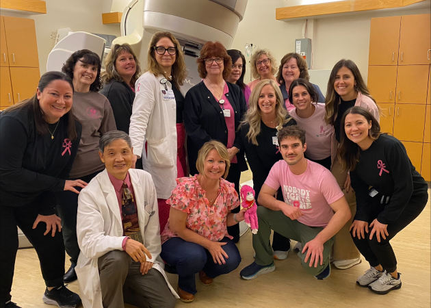 A group of medical professionals and staff, some wearing pink for breast cancer awareness, pose and smile together in a treatment room with medical equipment in the background.
