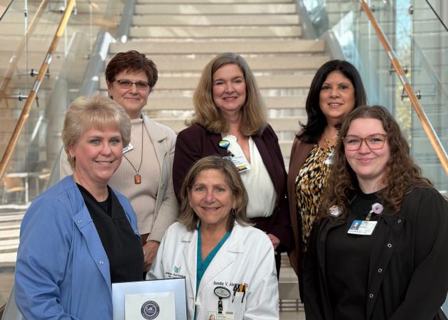 Six professional women pose together on a staircase, some wearing medical attire with name badges, while others wear business clothing, suggesting a healthcare or hospital setting. They are smiling and looking at the camera.