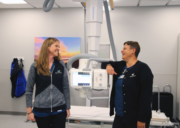 Two smiling healthcare workers stand next to an X-ray machine in a bright medical room, facing each other and laughing. Medical equipment and a nature photo are visible in the background.