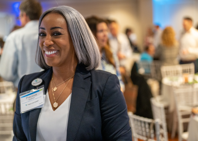 A woman with gray hair, wearing a navy blazer and name badge, smiles at an indoor event. Other people are visible in the blurred background, sitting and standing near white chairs.