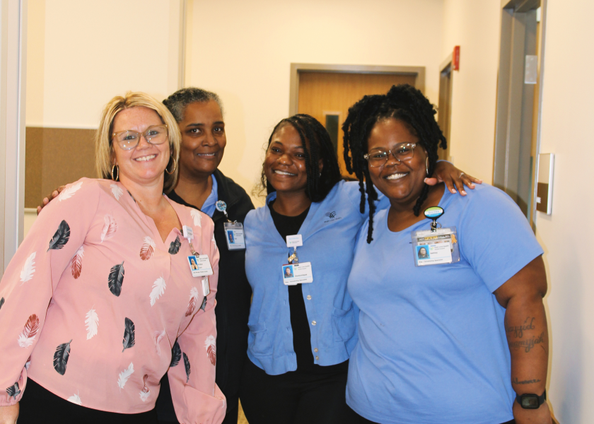 Four women, all wearing work badges and smiling, stand closely together in a hallway. Three wear blue, one wears a pink feather-patterned blouse. They appear happy and friendly in a professional setting.