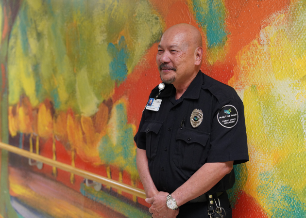A security guard in uniform stands and smiles in front of a colorful, abstract mural with shades of green, orange, and yellow.